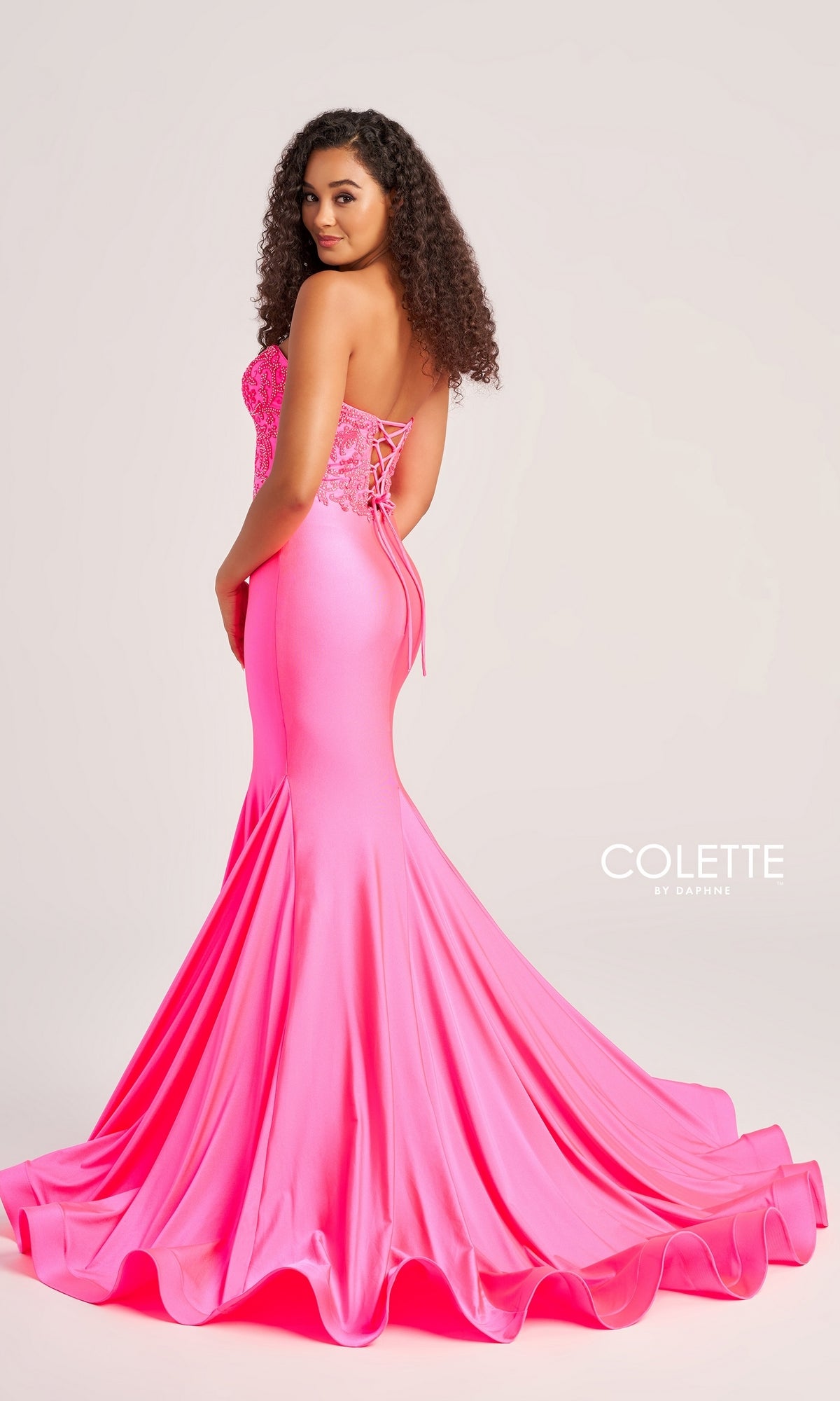 A woman in a bright pink Colette Strapless Long Mermaid Prom Dress CL5112 with lace-up back detail stands against a plain light background.